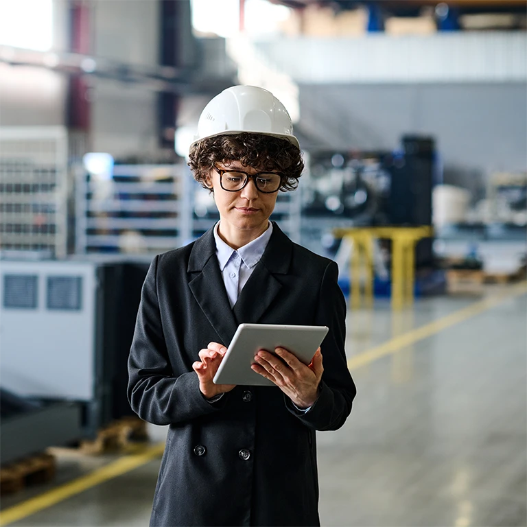 Woman walking in the warehouse with hardhat, taking notes on a touch pad.